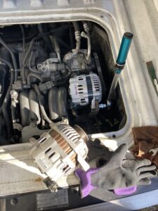 スバルサンバーのエンジンルーム内、オルタネーターを交換している画像。手前に古い部品があり、新たにセットされた部品をトルクレンチで固定ているところ。